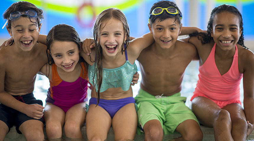 Diverse children smiling, sitting on the edge of a swimming pool.